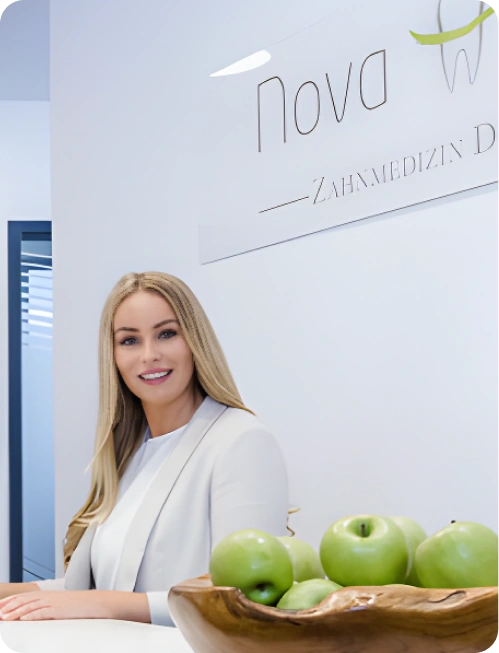 Nova-Smile Empfangsbereich mit freundlicher Mitarbeiterin am Empfang, Logo Nova-Smile im Hintergrund, grüne Äpfel in einer Holzschale, helle weiße Innenausstattung